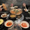 Una parrilla coreana de carbon al centro de la mesa, diversos platillos de opciones para asar alrededor, manos sosteniendo palillos chinos tomando comida
