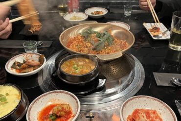 Una parrilla coreana de carbon al centro de la mesa, diversos platillos de opciones para asar alrededor, manos sosteniendo palillos chinos tomando comida