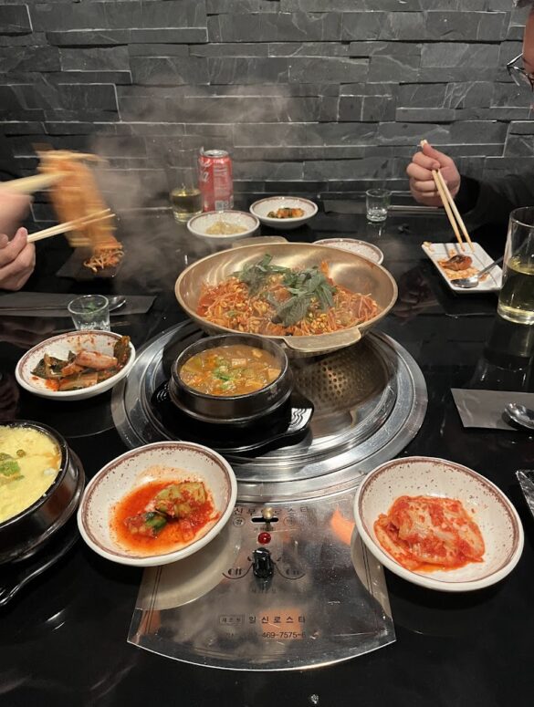 Una parrilla coreana de carbon al centro de la mesa, diversos platillos de opciones para asar alrededor, manos sosteniendo palillos chinos tomando comida